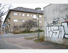 Straßenszene mit einem Wohngebäude aus beige-braunem Mauerwerk, mehreren Fenstern und kleinen Balkonen. Im Vordergrund eine mit Graffiti besprayte Wand, ein Gehweg aus Pflastersteinen, ein Straßenlaternenmast und ein karger Baum.