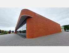 Moderne Backsteinfassade mit abgerundeten Ecken und einer langen Glasfront auf einer gepflasterten Fläche; Umgebung mit Bäumen und mehreren geparkten Autos im Hintergrund.