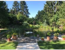 Ein gepflegter Garten mit einem kleinen Teich, umgeben von üppiger Vegetation und hohen Bäumen im Hintergrund. Im Vordergrund stehen ein runder Tisch mit Mosaikmuster, zwei Metallstühle und mehrere Blumentöpfe auf einer Holzterrasse.