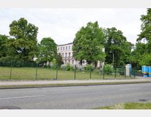 Ein mehrstöckiges weißes Gebäude mit klassizistischen Elementen, umgeben von einem großen, eingezäunten Garten mit Bäumen und Sträuchern. Im Vordergrund verlaufen ein Bürgersteig und eine Straße, im Hintergrund befinden sich weitere Grünflächen.