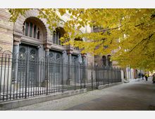 Historische Gebäudefassade aus gelbem und rotem Backstein mit kunstvoll verzierten, hohen Eingangstüren aus Metall hinter einem schwarzen Zaun; daneben Gehweg mit gelb gefärbten Baumkronen im Herbst.