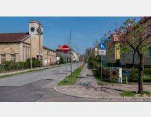 Straßenszene in einer Wohngegend mit Gehweg und Fahrbahn, flankiert von Reihenhäusern mit gelben und braunen Fassaden. Ein markantes Gebäude mit Uhrturm aus Backstein befindet sich links, während blühende Bäume und Verkehrsschilder den Gehweg auf der rechten Seite säumen.
