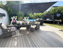 Große Holzterrasse im Garten mit Rattan-Loungegruppe und Esstisch unter einem breiten Sonnenschirm. Im Hintergrund stehen ein aufgestellter Pool, ein abgedeckter Grill sowie ein Trampolin und ein Basketballkorb vor hohen Hecken.