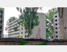 Mehrgeschossige verlassene Plattenbauten mit leeren Fensteröffnungen, umgeben von Bäumen. Im Vordergrund ein Stück einer Betonmauer mit vereinzeltem Graffiti.