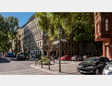 Straßenecke in einem urbanen Wohnviertel mit mehrstöckigen, hellen Altbauten, bepflanzten Balkonen und Bäumen, die Schatten spenden. Im Vordergrund Straßenschilder, parkende Autos und ein kleiner bepflanzter Platz.