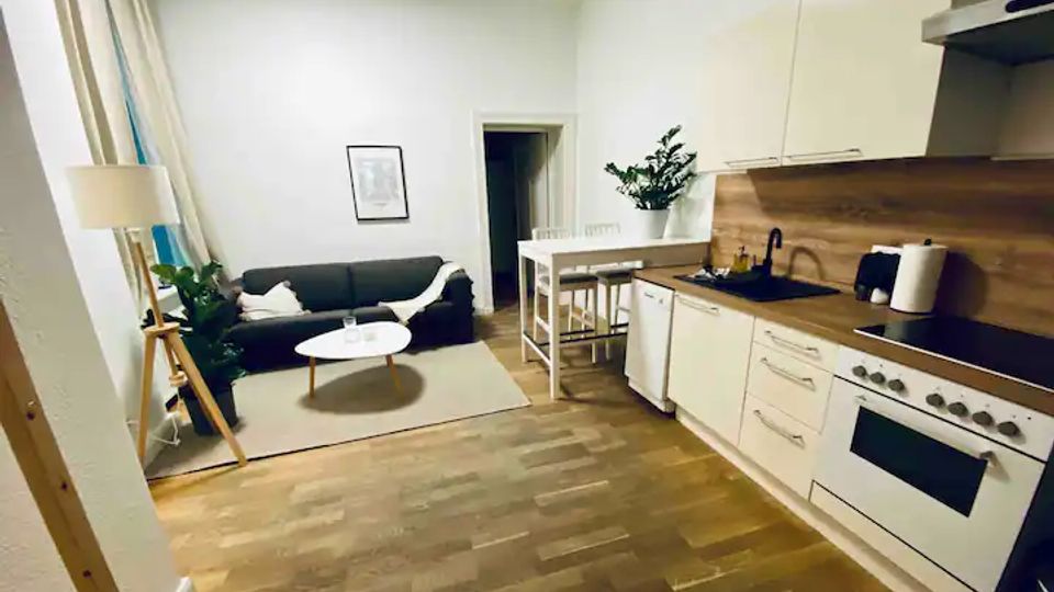 Offener Wohnbereich mit integrierter Küchenzeile in Weiß, Holzarbeitsplatte und dunkler Spüle sowie Herd und Backofen. Daneben stehen ein kleiner Esstisch mit Hockern und eine Sitzecke mit Sofa, rundem Couchtisch, Stehlampe und großem Fenster mit Vorhängen auf Holzboden.