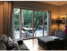 Wohnzimmer mit grauem Sofa und Zugang zu einer Terrasse durch eine große Glastür. Auf der Terrasse stehen ein Holztisch mit Bänken, umgeben von Pflanzen und Lichterketten, mit einem Blick auf einen Garten mit Bäumen.