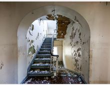 Blick in ein verfallenes Treppenhaus mit grauen Stufen und Geländer unter einem Rundbogen. An den Wänden und der Decke blättert großflächig die Farbe ab, und am oberen Ende fällt Licht durch ein Fenster.