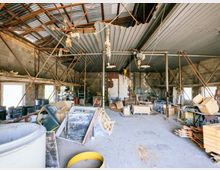 Ein altes, heruntergekommenes Lagerhaus mit Wellblechdach, freiliegenden tragenden Metallstreben und unverputzten Betonwänden. Der Raum ist gefüllt mit verschiedenen Materialien, Kartons, Werkzeugen und verstreuten Gegenständen unter einem teilweise beschädigten Dach.