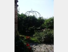 Ein kleiner Garten mit Kopfsteinpflaster, umgeben von dichtem Grün, roten und weißen Blumen. In der Mitte steht eine filigrane Metallpergola mit einer Sitzgruppe im Schatten.