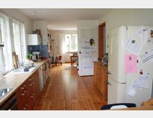 Helle Küche mit langen Holzarbeitsplatten, weißen Wänden und Holzboden. Rechts steht ein cremefarbener Retro-Kühlschrank, an dem Zettel und Magnete hängen, während eine Tür mit Kinderzeichnungen zum Essbereich mit Fenster führt.