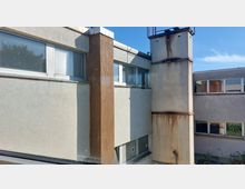 Außenansicht eines Gebäudekomplexes mit flachen Dächern und grober Putzfassade. Auf der rechten Seite ist ein Industrie-Schornstein mit Rostspuren zu sehen, links und rechts Fenster mit Jalousien.