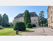 Ein historisches Gebäude mit heller Fassade und dunklem Schrägdach, umgeben von gepflegtem Rasen, geschnittenen Sträuchern und hohen Bäumen. Im Vordergrund ist ein gepflasterter Weg mit einem Mülleimer zu sehen.