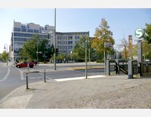 Straßenszene in einer städtischen Umgebung mit modernem Gebäude im Hintergrund, einem S-Bahnhof-Eingang auf der rechten Seite und Verkehrsampeln sowie Gehwegen im Vordergrund. Bäume und vereinzelte Fahrzeuge ergänzen die Umgebung.