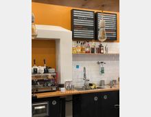 Ein Teil eines modernen Cafés mit einer hellen Theke aus Holz, einer Espressomaschine und gestapelten Tassen. Im Hintergrund sind weiße Kacheln, ein Waschbecken mit einer hohen Armatur, sowie ein Regal mit Getränkeflaschen und ein Menübrett an einer orangefarbenen Wand zu sehen.