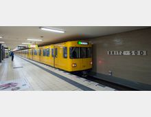 Untergrund-U-Bahn-Station „Britz-Süd“ mit einem gelben U-Bahn-Wagen, der am Bahnsteig steht. Die Umgebung besteht aus beigen Wandfliesen, hellem Boden und Leuchtschildern für Orientierung.