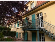 Gelbes Wohngebäude mit roten Dachziegeln, türkisfarbenen Fensterläden und einem kleinen Balkon mit Staubenkonstruktion. Im Vordergrund befinden sich ein Baum mit dunkelrotem Laub sowie ein mit Pflanzen umgebenes Sitzbereich mit Holzmöbeln.
