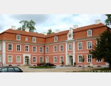 Rosa Herrenhaus mit weiß abgesetzten Fenstern und Türen, rotem Mansardendach und einer Uhr über dem Haupteingang. Im Vordergrund ein gepflasterter Hof mit parkenden Autos und kleinen Grünflächen.