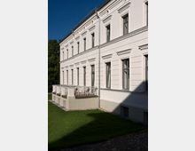 Seitenansicht eines weißen, zweigeschossigen Gebäudes mit symmetrischen Fenstern und klassizistischen Stilelementen. Im Vordergrund eine kleine Terrasse mit weißen Metallgeländern und Gartenmöbeln, umgeben von einer Rasenfläche.