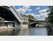 Eine Brücke mit einem gelben Bus darauf überquert einen Fluss, flankiert von einem Gebäude mit moderner Treppenarchitektur und Fassaden mit Graffiti. Im Hintergrund sind Bäume und blauer Himmel mit wenigen Wolken zu sehen.