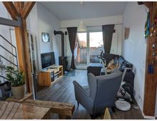 Wohnzimmer in einer modernen Wohnung mit Holzbalken, einer grauen Couch, einem Fernseher auf einem Holzmöbelstück und einem Kaminofen. Im Hintergrund befinden sich eine große Glas-Schiebetür mit Zugang zu einem Balkon und dunklen Vorhängen.