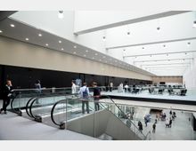 Offenes, modernes Atrium in einem öffentlichen Gebäude mit hell beleuchteter Decke, Glasgeländern und Rolltreppen, die zu mehreren Ebenen führen. Menschen gehen und sitzen in verschiedenen Bereichen, darunter Wartezonen mit Stühlen und Tischen.