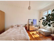 Helles Schlafzimmer mit Holzboden und hohen Decken, eingerichtet mit einem Bett, einem kleinen Nachttisch, einer Metallkommode, einem Glassofatisch und einem weißen Sofa. Große Fenster mit Vorhängen lassen natürliches Licht herein; eine Zimmerpflanze steht rechts im Vordergrund.