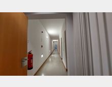 Langer, schlichter Innenflur mit weißen Wänden und hellem, glänzendem Boden. An der linken Wand befinden sich ein Feuerlöscher sowie mehrere Wandlampen und kleine Lichtquellen in Bodennähe; Türen verlaufen beidseitig entlang des Korridors.