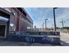 Außenbereich eines S-Bahnhofs mit moderner Metall- und Glasfassade, teilweise mit Graffiti bedeckt. Im Hintergrund sind Gleise, eine Brücke und eine städtische Skyline mit Hochhäusern zu sehen.