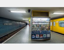 U-Bahn-Station "Parchimer Allee" in Berlin mit einem gelben Zug auf der rechten Seite, einer Kioskvitrine mit Getränken und Zeitschriften in der Mitte sowie einem mit blauen Fliesen verkleideten Bahnsteig auf der linken Seite. Die beleuchtete Station hat eine gerade Anordnung mit Schienen, die in die Ferne führen.