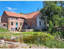 Backsteingebäude mit rotem Ziegeldach und blauen Türen, umgeben von einer grünen Landschaft. Im Vordergrund befindet sich ein kleiner Teich mit Pflanzen und eine hölzerne Begrenzung, umgeben von Steinen und Wiesen.