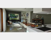 Moderne Wohnküche mit Essbereich, einem großen Holztisch und weißen Stühlen. Im Hintergrund ein Flügel, ein großes Fenster mit Blick in den Garten und dekorative Elemente an den Wänden; im Vordergrund eine Küchenzeile mit einer Schale Obst.