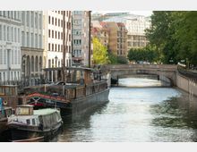 Stadtkanal mit festgemachten Schiffen und Gebäuden im Hintergrund, flankiert von einer steinernen Brücke und Bäumen. Die Umgebung zeigt dicht bebaute, mehrstöckige Häuser in urbaner Umgebung.