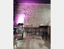 Ein Innenraum mit einer freiliegenden Ziegelwand, die von einer rosa Beleuchtung erhellt wird. Vor der Wand stehen hohe Tische mit Hockern, während im Vordergrund mehrere Sessel und kleine runde Holztische auf einem Holzfußboden angeordnet sind.