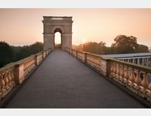 Arkadenbrücke mit historischen Geländern, die zu einem hohen Torbogen führt, im Hintergrund Bäume und ein sanfter Sonnenaufgang am Himmel. Die Umgebung wirkt ruhig und von klassischer Architektur geprägt.