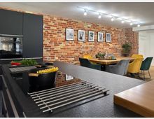 Moderne Wohnküche mit einer Kochinsel aus dunklem Stein, darauf zwei Kochtöpfe sowie frische Zutaten. Im Hintergrund eine Ziegelsteinwand mit schwarzen Bilderrahmen und ein Essbereich mit Holztisch und bunten Samtstühlen.