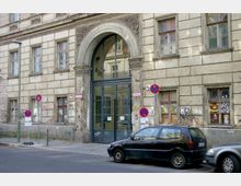 Historische Hausfassade mit sichtbaren Abnutzungsspuren, mehreren Fenstern sowie einem großen, verzierten Rundbogenportal mit Glas- und Metalleinsatz. Im Vordergrund stehen Autos am Straßenrand, und Parkverbotsschilder sind deutlich sichtbar.