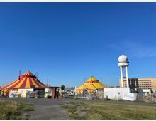 Zwei bunte Zirkuszelte in Rot-Gelb und Gelb-Rot stehen auf einer Wiese, umgeben von Zäunen und Baucontainern. Im Hintergrund befinden sich ein weißer Wasserturm mit Kuppel sowie ein großes, rechteckiges Gebäude, alles unter einem wolkenlosen blauen Himmel.