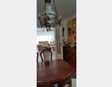 Esszimmer mit Holztisch und Stühlen im Vordergrund, einem dekorativen Kronleuchter an der Decke und einer halb geöffneten Tür, die in ein Wohnzimmer mit Sofas und großen Fenstern führt. An der Wand hängen gerahmte Bilder, und eine Kommode mit Lampen und Dekorationen steht rechts.