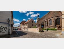 Straßenszene in einer historischen Altstadt mit gepflasterten Straßen. Auf der rechten Seite steht eine große Kirche aus Backstein mit hohen Fenstern, links befinden sich Reihen von hellen Wohnhäusern mit roten Dächern.