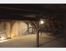 Dunkler Dachbodenraum mit sichtbaren Holzbalken, alten Holzdielen und einer unverputzten Ziegelwand. Licht fällt durch ein kleines Fenster in der Dachschräge, das sich zwischen den Holzverstrebungen befindet.