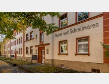 Fassade eines Gebäudes mit der Aufschrift "Papier und Schreibwaren", helle Putzwände mit rot-braunen Fensterrahmen und Türumrandungen, umgeben von Bäumen mit herbstlichem Laub. Der Gehweg davor ist teils von Laub bedeckt.
