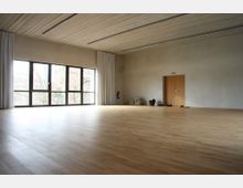 Ein leerer Raum mit hellem Holzboden, großen bodentiefen Fenstern mit weißen Vorhängen auf der linken Seite und einer Tür mit Holztäfelung an der rechten Wand. In der Ecke liegen gefaltete Matten und einige Schuhe.