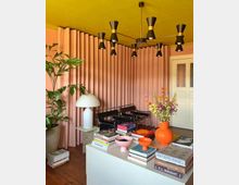 Wohnzimmer mit pastellfarbenem Interieur: Rosa Vorhänge bedecken eine Wand, ein senfgelber Deckenleuchter mit schwarzen Elementen hängt zentral, und ein weißer Tisch mit bunten Dekorationen und Büchern steht im Vordergrund. Zwei schwarze Stühle, eine Topfpflanze und eine weiße Tischlampe ergänzen den Raum.