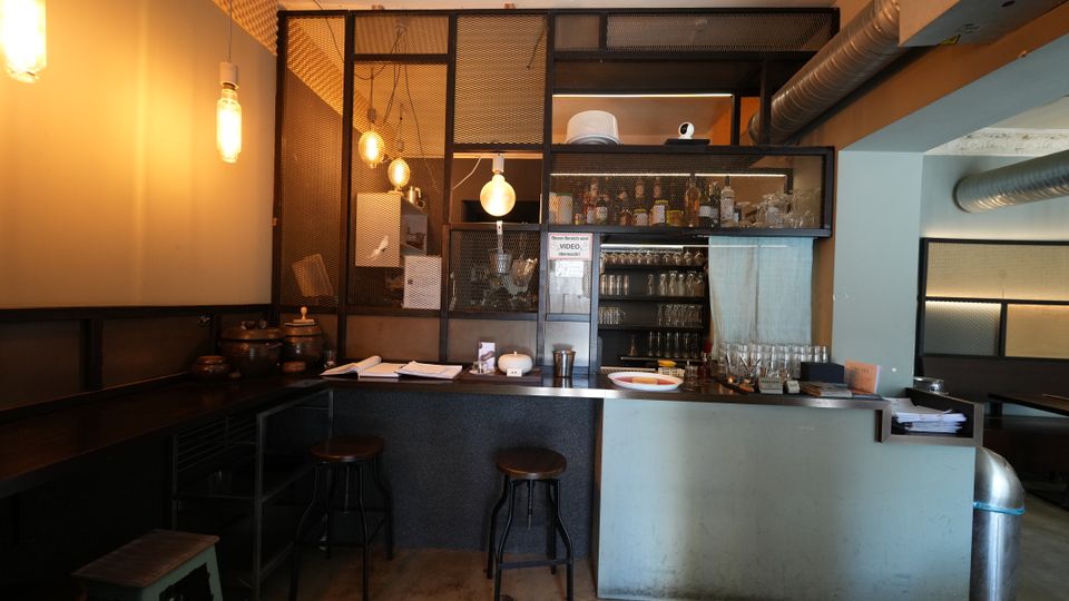 Innenraum einer Bar mit langem Tresen, Barhockern und Regalflächen mit Flaschen und Gläsern hinter einem Metallgitter. Warmes Licht durch hängende Glühbirnen, dazu sichtbare Lüftungsrohre und industrielle Oberflächen wie Betonboden und dunkle Wandverkleidung.