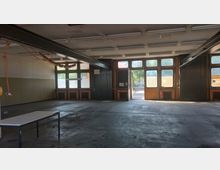 Ein leerer Industriebereich mit großflächigen Fenstern und Türen aus Glas und Metall. Der Betonboden, sichtbare Deckenverstrebungen, hängende Kabel und ein einzelner Tisch betonen die funktionale Umgebung.