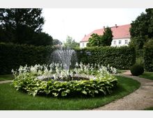Brunnen in einer Gartenanlage mit kreisförmigem Becken, umgeben von blühenden weißen Blumen und grünem Laub. Im Hintergrund Hecken, Bäume und ein Gebäude mit rotem Ziegeldach.