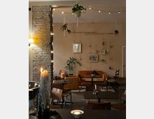 Gemütliches Café mit dunklen Holztischen und Stühlen, orangefarbenem Sofa im Hintergrund und dekorativer Beleuchtung durch Lichterketten. An der hellen Wand hängen kleine Pflanzen, Bilder und Spiegel; ein Kerzenhalter mit tropfendem Wachs steht im Vordergrund.