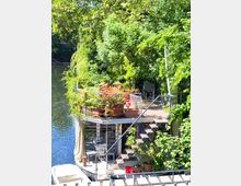 Hausboot mit zwei Ebenen, umgeben von dichtem Grün direkt am Wasser. Die obere Terrasse ist mit Pflanzen in Blumentöpfen gestaltet, und es stehen rote Stühle sowie weitere Sitzmöglichkeiten bereit; im unteren Bereich befinden sich eine überdachte Sitzgruppe und eine Treppe, die beide Ebenen verbindet.