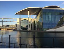 Moderne Architektur am Spreeufer in Berlin, mit einem großen Gebäude aus Glas und Beton, das durch eine markante, runde Fensteröffnung und ein geschwungenes Dach auffällt. Im Vordergrund fließt die Spree, und eine Fußgängerbrücke verbindet die Gebäude.